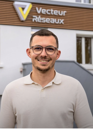 Smiling man wearing glasses and a light polo, standing outside a building with a Vecteur Réseaux sign.