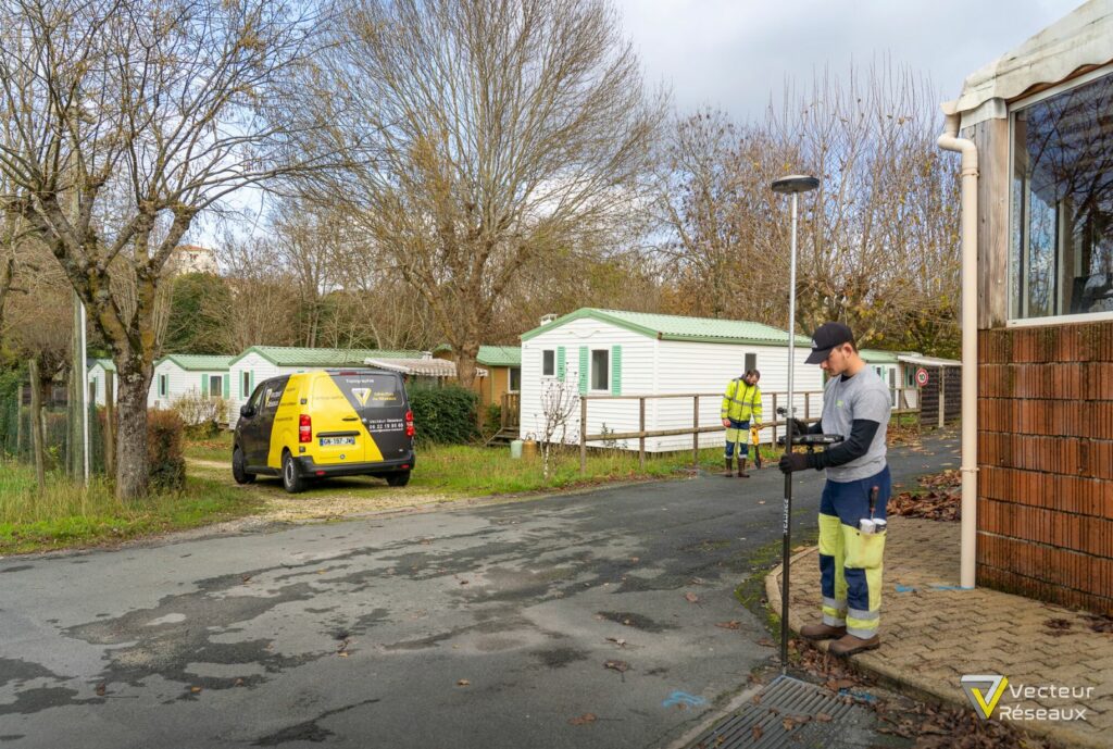 Two workers in high‑visibility clothing near a yellow service van in a mobile home park, with leafless trees and white modular homes in the background.
