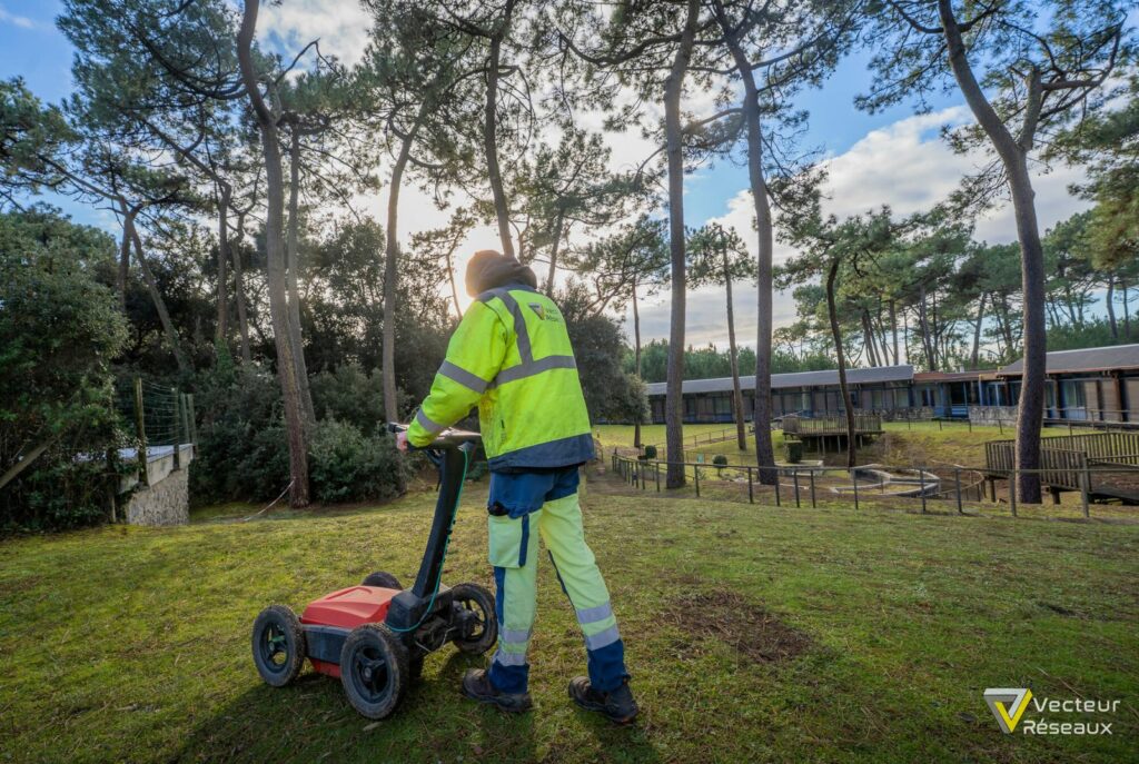 Utility worker in a high-visibility jacket operating a red ride-on lawn mower on a grassy area among tall pine trees, daylight.
