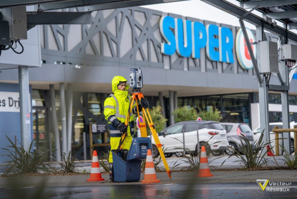 Surveyor in neon rain gear operating a surveying instrument on a tripod outside a shopping center with a 'SUPER U' sign in the background.