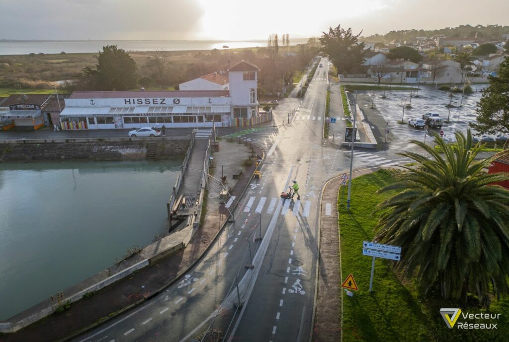 Aerial view of a seaside town with a canal, a white building labeled 'HISSEZ Ø', and a person riding a small street-cleaning vehicle on a crosswalk.
