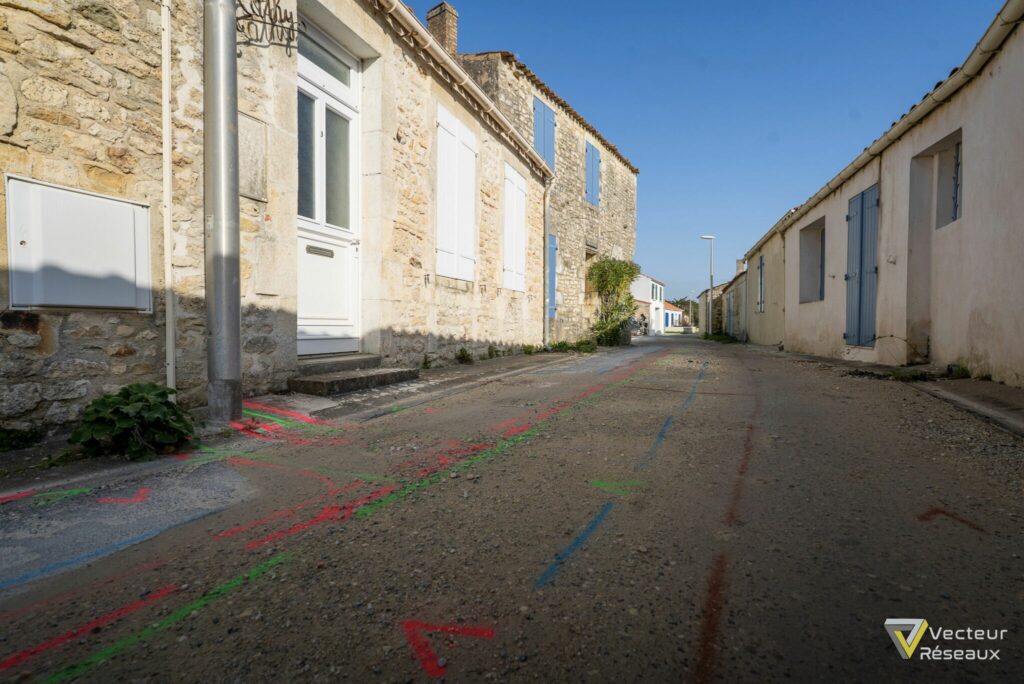 Narrow sunlit street in a stone village with white doors and blue shutters, colorful chalk markings on the pavement, clear blue sky above.