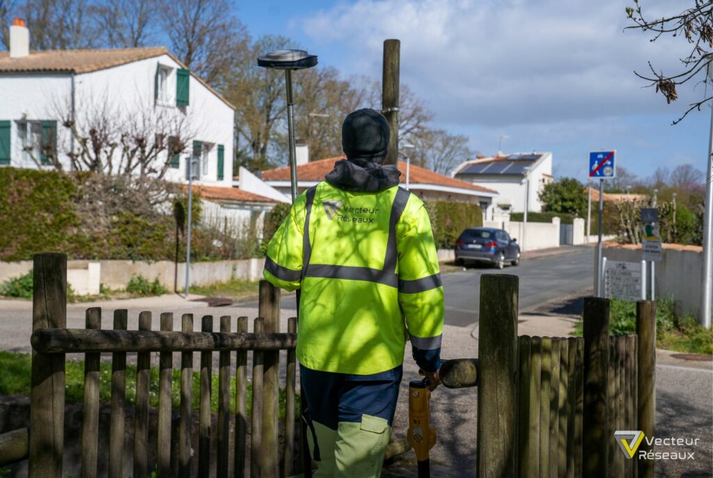 Utility worker in a neon safety jacket with 'Vecteur Réseaux' on the back stands at a wooden gate on a residential street, holding a tool.
