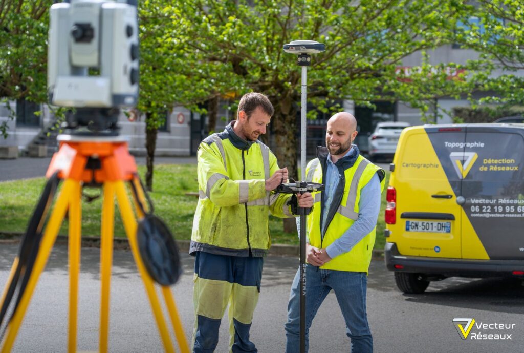 Two men in high-visibility jackets set up surveying equipment outdoors, adjusting a total station mounted on a pole with a tripod nearby, in front of a yellow work van.