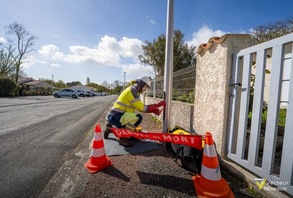 Worker in high-visibility jacket and helmet repairing or inspecting a streetlight next to a gated yard, with orange safety cones and 'DANGER DE MORT' tape nearby